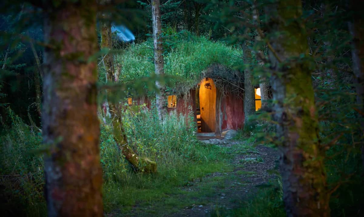 Magical Woodland Glamping North Wales Hobbit House view of the front through the trees and the lights on inside the earth house