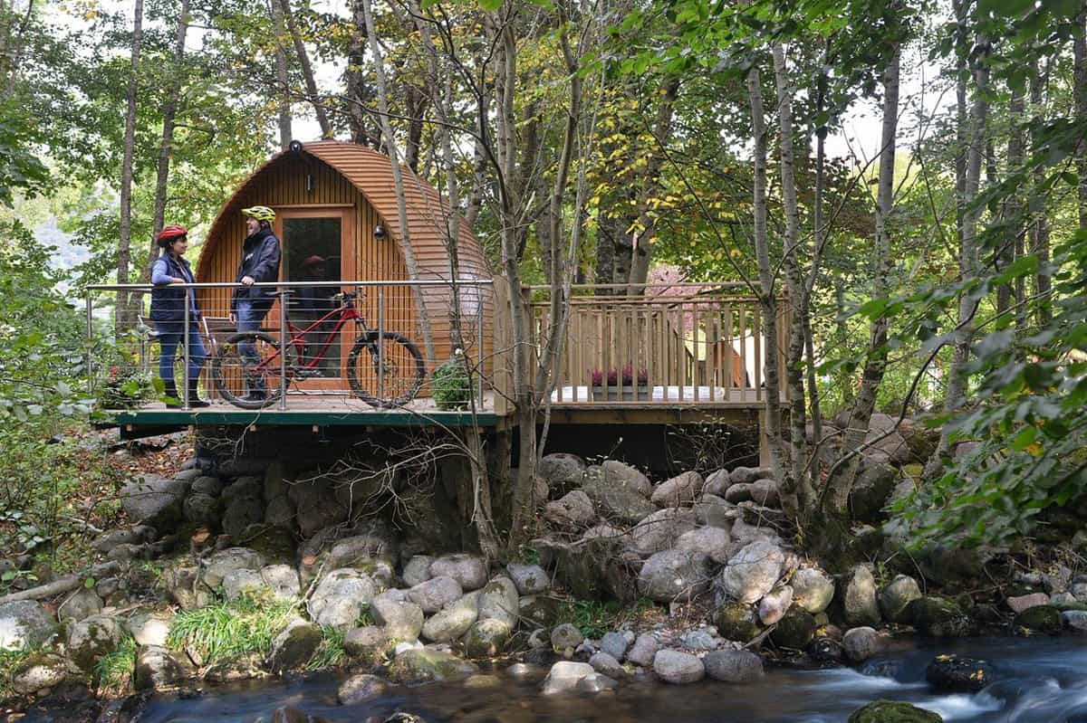 All About Pod Glamping featured image glamping pod with deck above a creek with cyclists on the deck