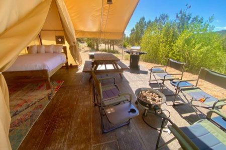view from east zion glamping tent deck with door open and bed inside and chairs, picnic table, bbq and firepit on the deck