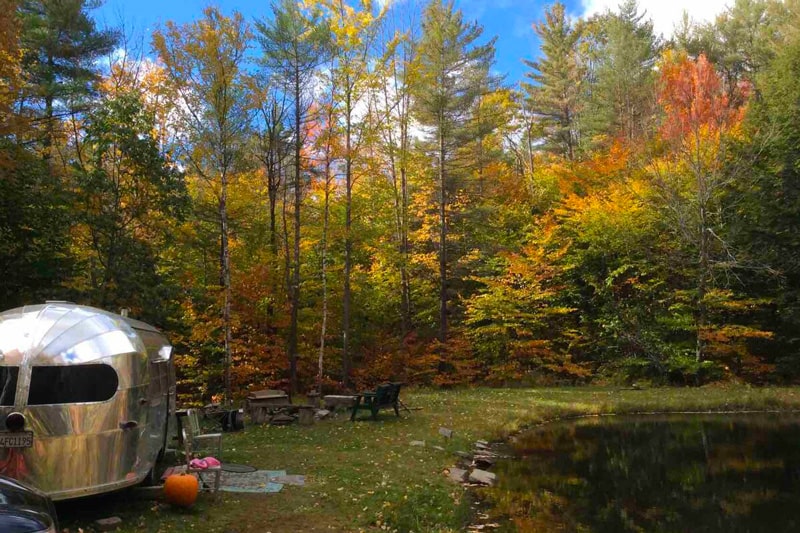Good Night Moon Vintage Airstream Glamping Vermont view of trailer, campfire and pond with colorful trees behind