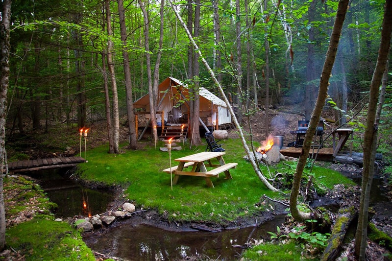 Hemlock Camp Glamping in the Adirondacks view of the camp with stream, picnic table, firepit with fire, bbq and glamping tent with tiki torches