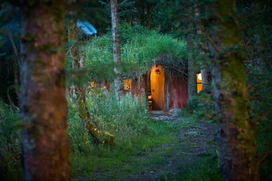 Magical Woodland Glamping Hobbit House view of the front through the trees and the lights on inside the earth house