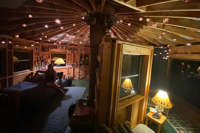 view of inside of treehouse at night with bookshelves and a person lounging on sofa
