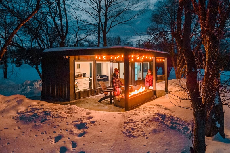 The Scenic Orchard Glamping in the Adirondacks in Winter with a view at night of the tiny home with the long fireplace outside burning and two people standing next to it
