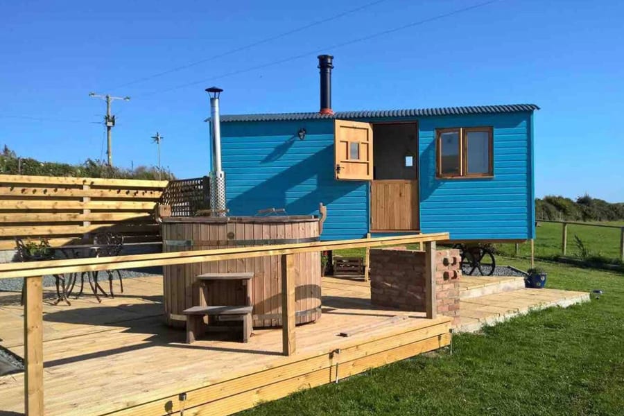 Isle of Anglesey Glamping with Hot Tub view of the blue shepherds hut with top half of door open and a deck with wooden hot tub on it