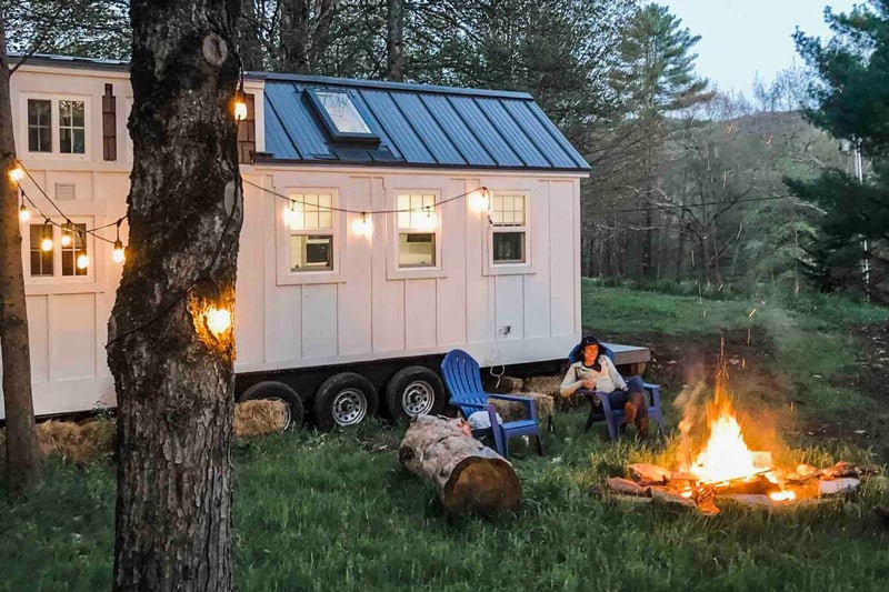 The Green Mountain Tiny House Vermont view of the side of white tiny home with campfire and chairs with one person next to fire